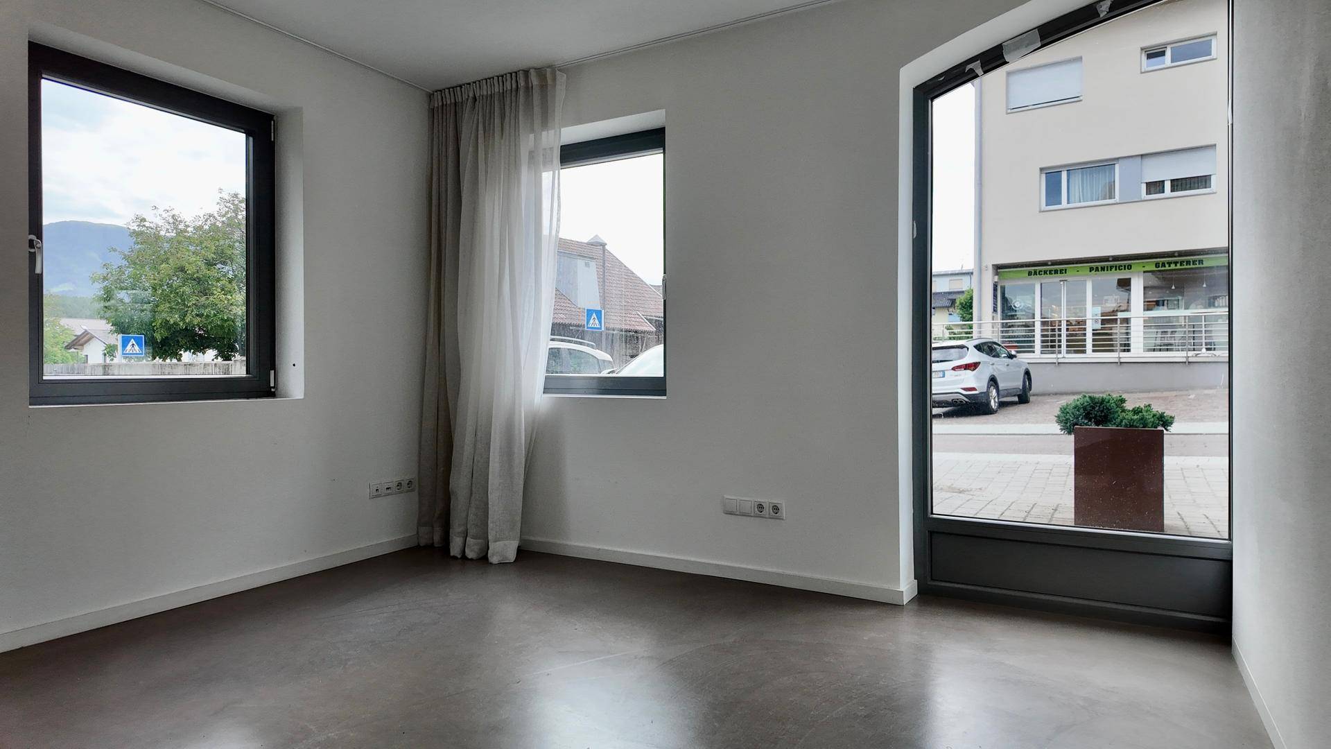 Heller Raum mit zwei Fenstern und einer großen Glastür, die den Blick auf die Straße freigibt.
