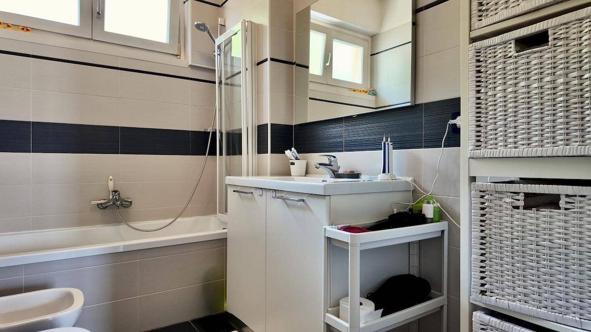 Badezimmer mit Waschtisch, Spiegel, Badewanne und Duschvorrichtung, in hellen und dunklen Fliesen.