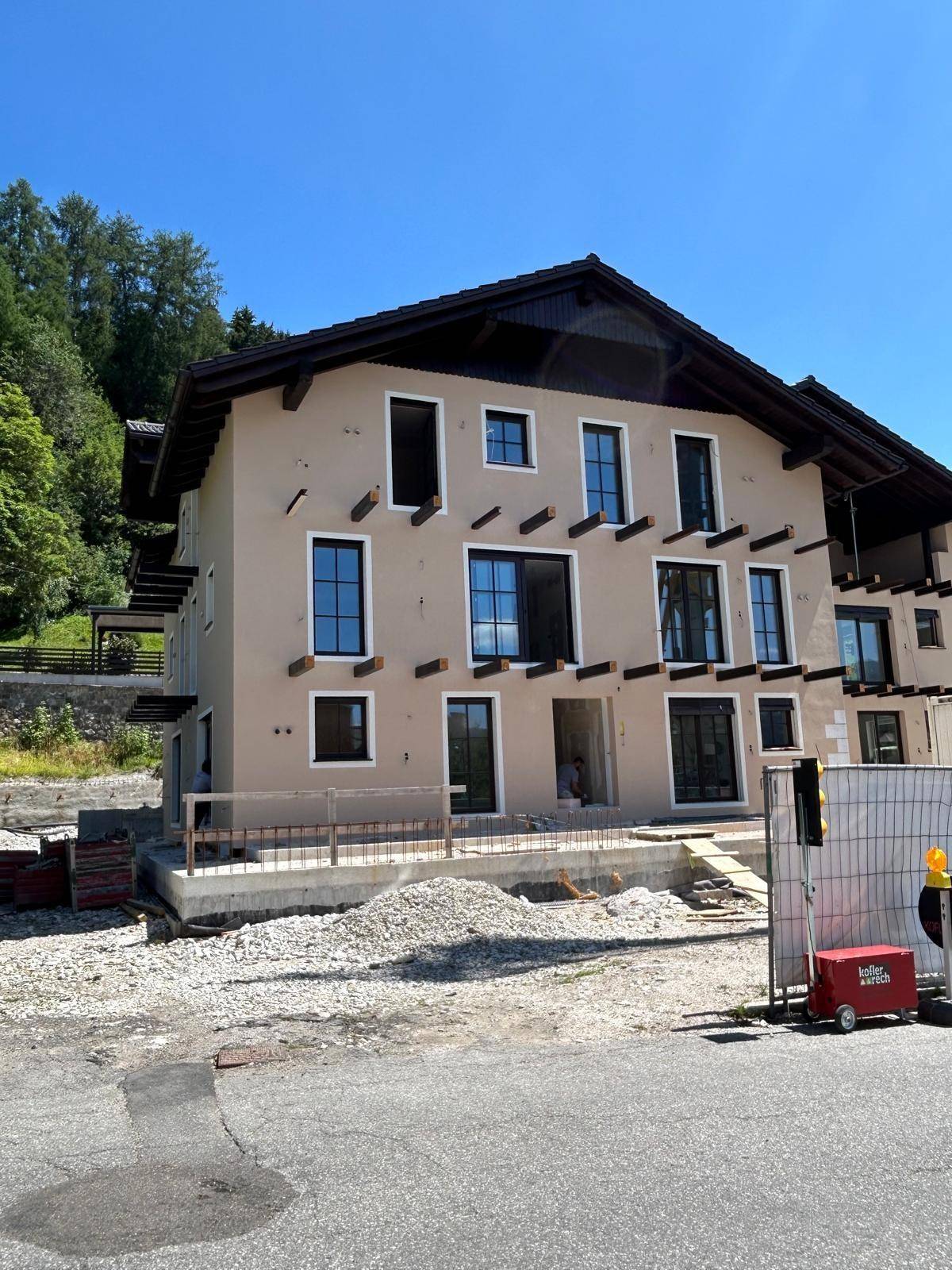 Vorderansicht des Neubaus mit beiger Fassade und großen Fenstern, umgeben von der Baustelle.