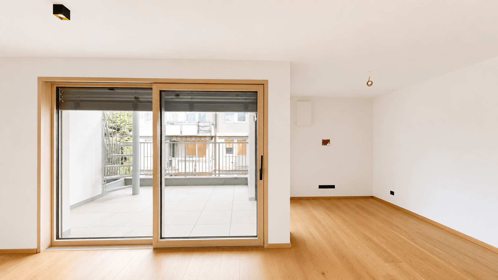 Leerer Wohnraum mit hellem Parkettboden und großem Schiebefenster zum Balkon.