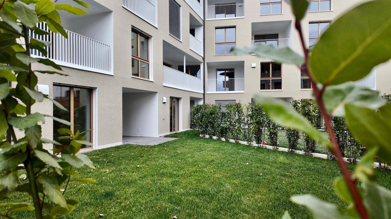 Grüner Rasenbereich mit Hecke vor einem modernen Wohngebäude mit Balkonen und Terrassenzugang.