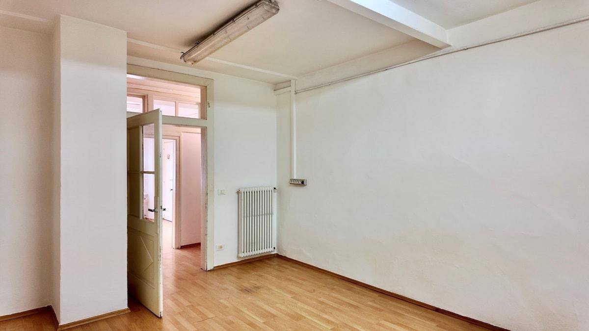 Heller, leerer Raum mit Holzfußboden, Heizkörper und offener Tür zu einem weiteren Zimmer.