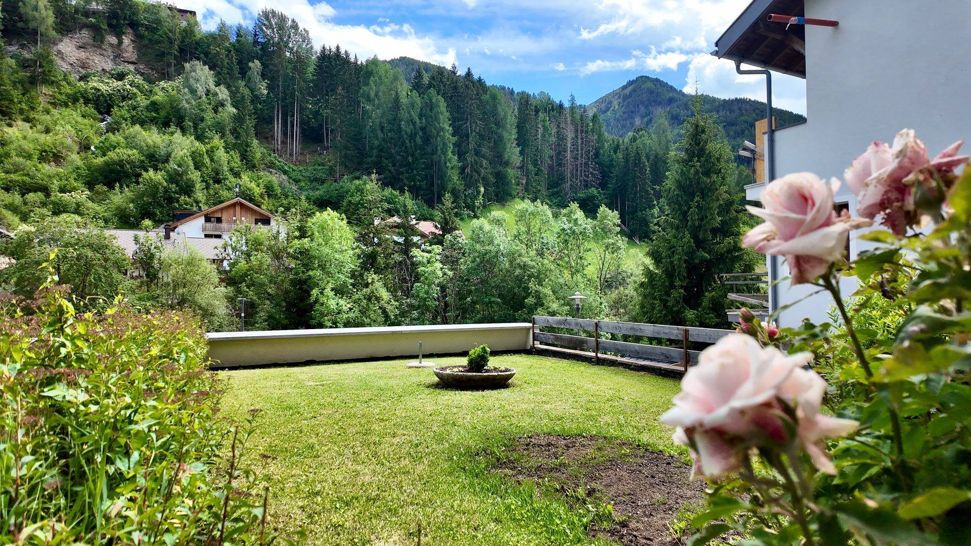 Gepflegter Garten mit Rasenfläche, Rosenbüschen und einem malerischen Blick auf die bewaldeten Berge.