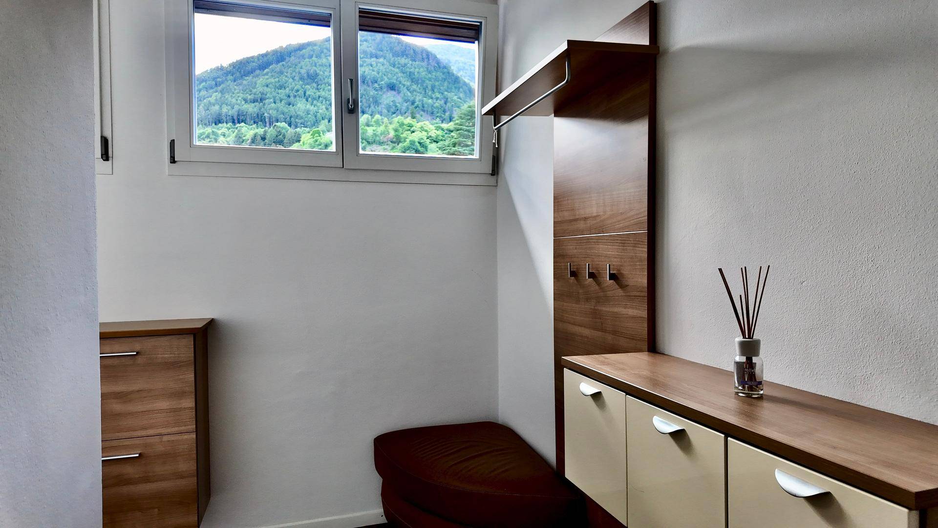 Heller Eingangsbereich mit Holzmöbeln, Garderobe und Fenster mit Bergblick.