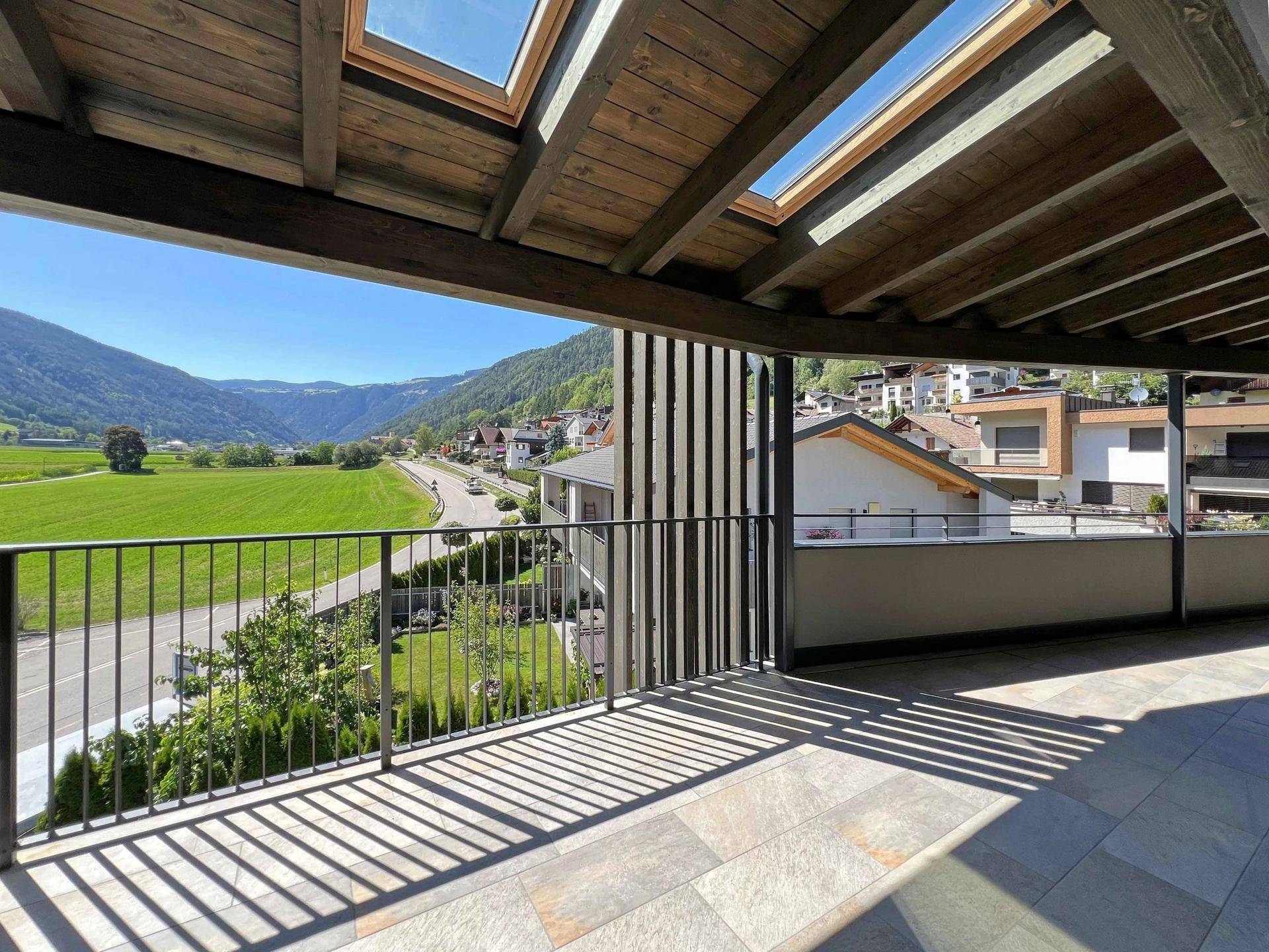 Helle Terrasse mit Holzbalkendecke, Dachfenstern und Ausblick auf die Landschaft und nahegelegene Häuser.
