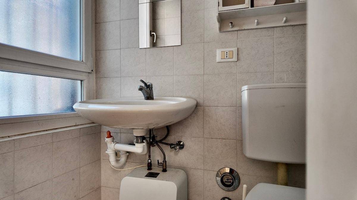 Badezimmer mit hellen Fliesen, Waschbecken, Toilette und einem Fenster für Tageslicht.