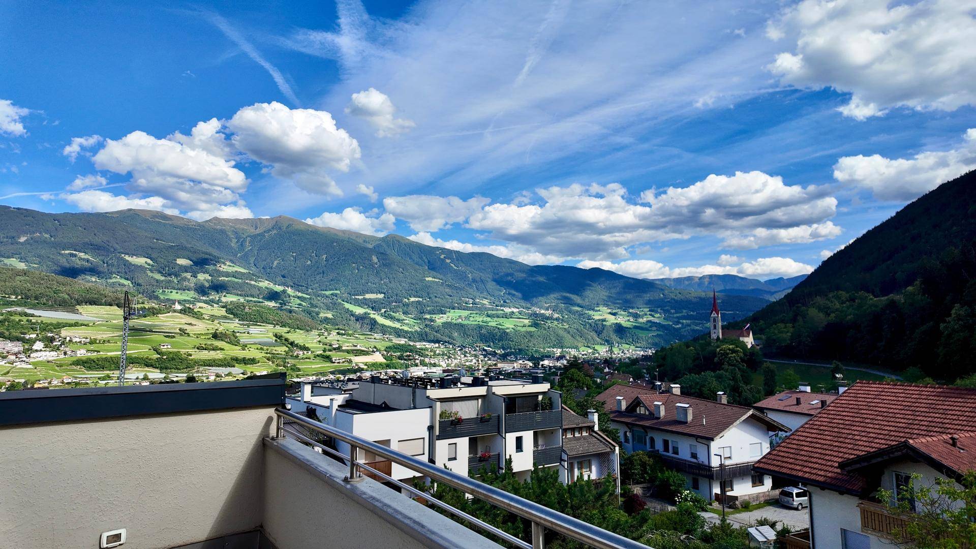 Weitläufiger Ausblick vom Balkon auf eine Stadt im Tal und die umliegenden Berge.