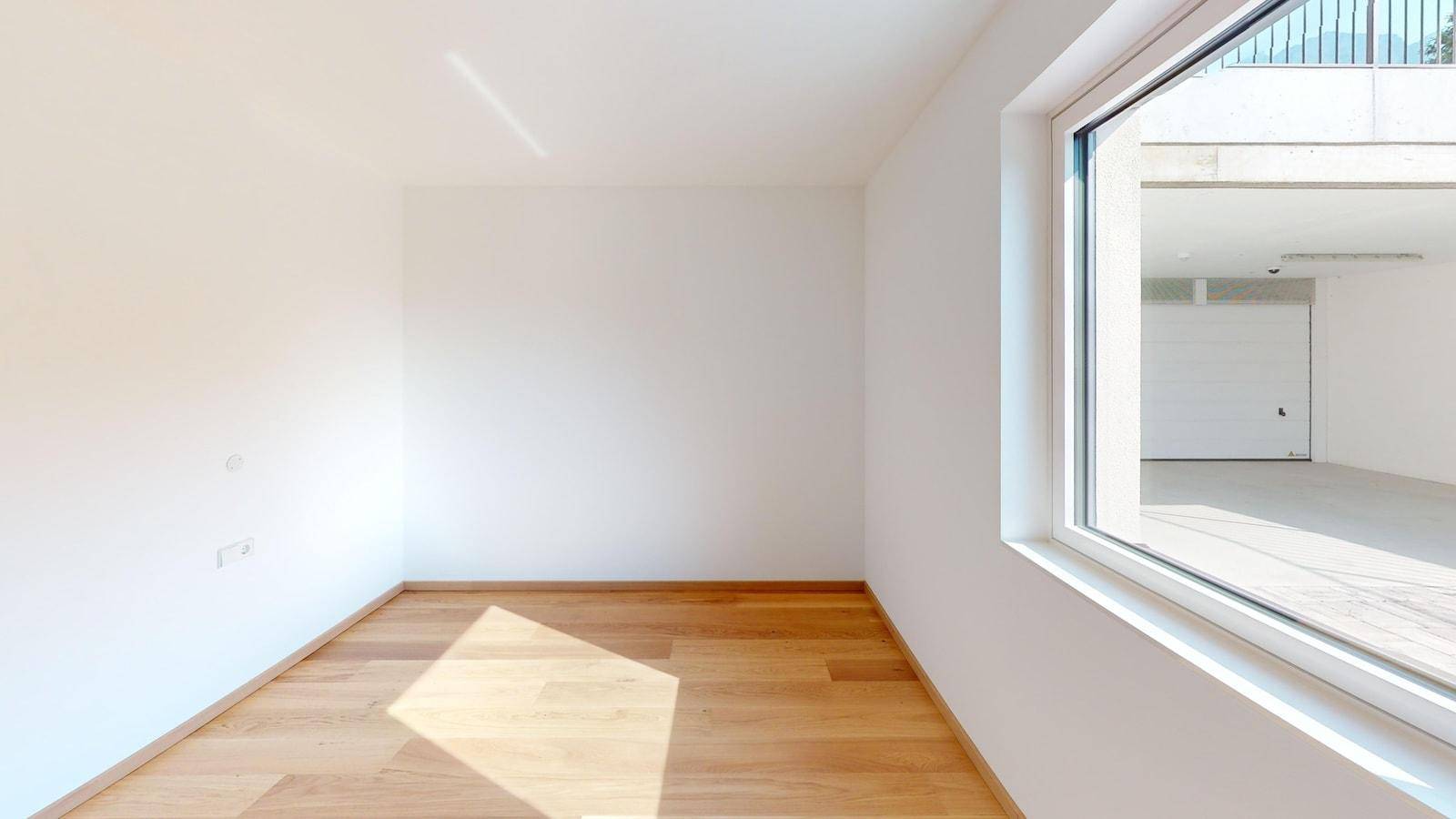Helles Zimmer mit Holzboden und Fenster, das einen Blick auf den Garagenbereich bietet.
