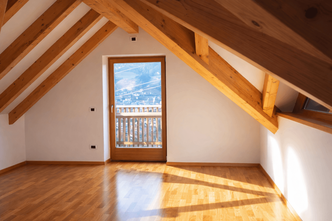 Leerer Raum mit Holzboden, Dachschräge, sichtbaren Holzbalken und Balkonzugang.