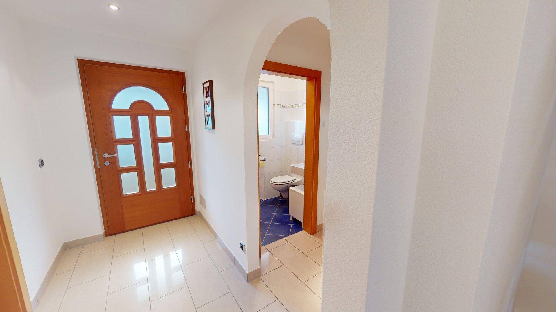 Heller Eingangsbereich mit Holztür und Blick in ein kleines Badezimmer mit WC.