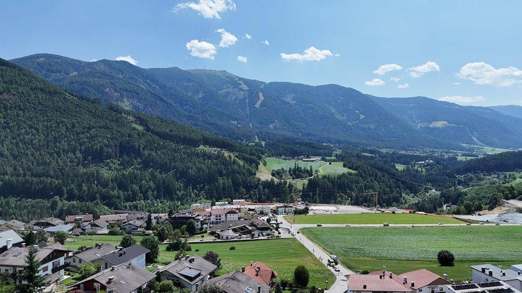 Weitläufige Ansicht eines Dorfes umgeben von grünen Bergen und weiten Feldern unter blauem Himmel.