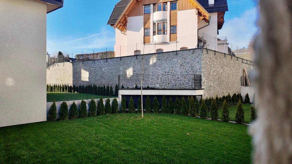 Gepflegter Gartenbereich mit Rasen und Hecken vor einer Steinmauer und Gebäuden.