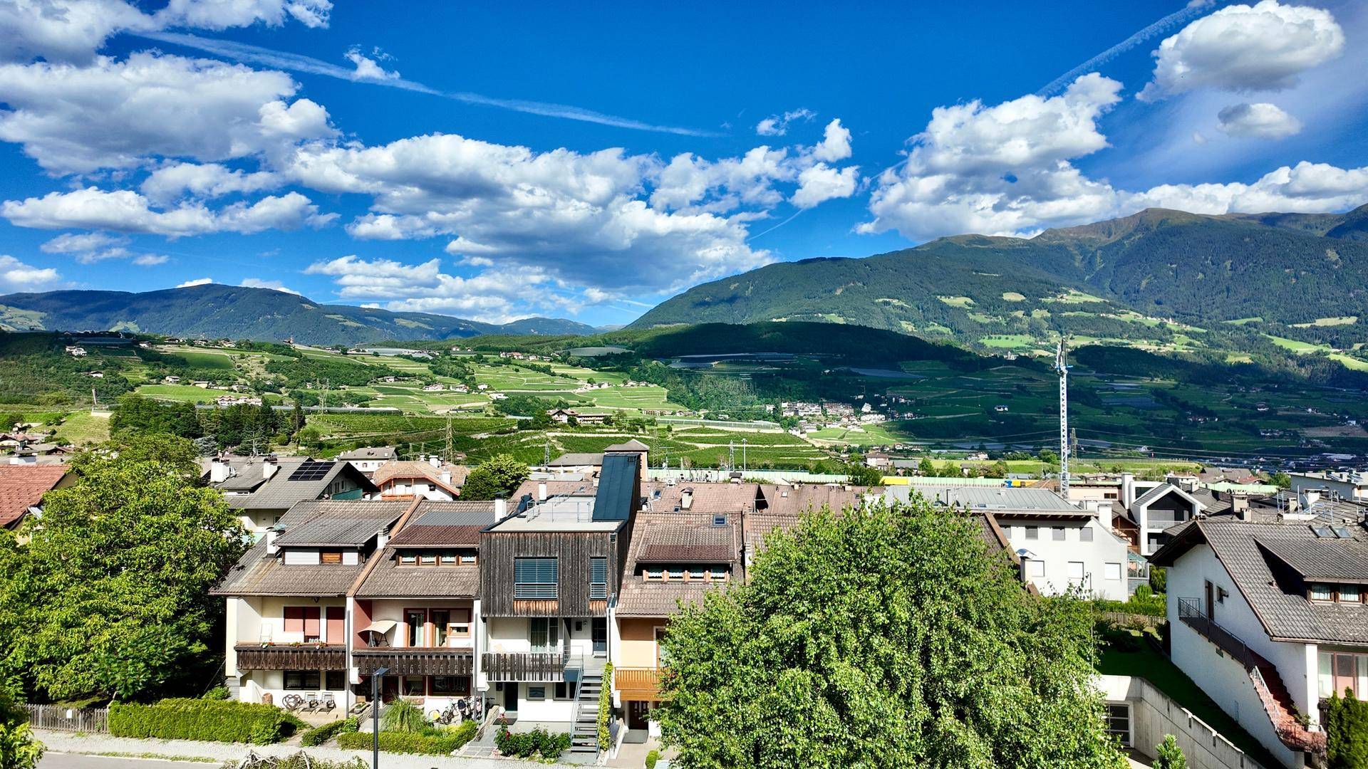 Panoramablick über ein grünes Tal mit Häusern und majestätischen Bergen unter blauem Himmel.