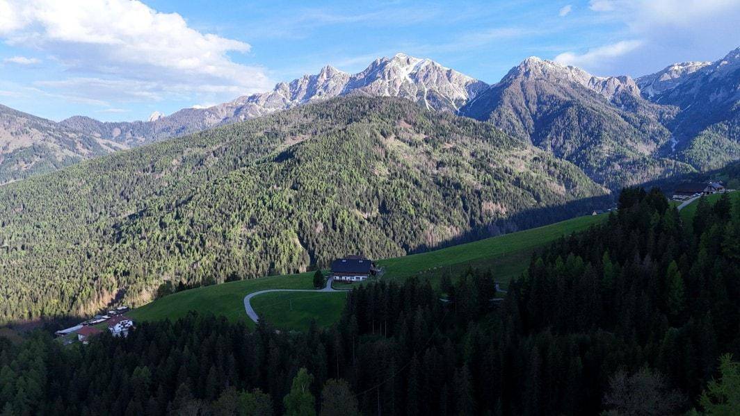 Weitläufige Berglandschaft mit dichten Wäldern und einem abgelegenen Haus in der Ferne unter blauem Himmel.