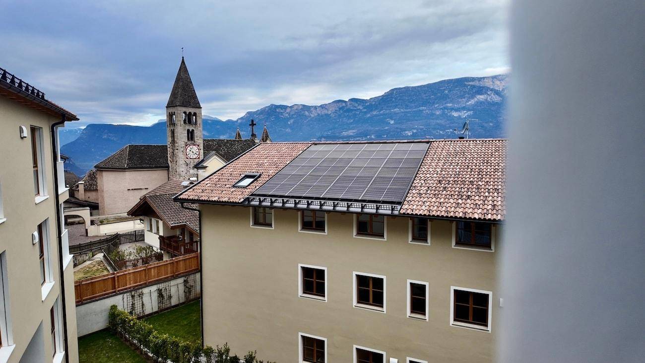 Gebäude mit Ziegeldach und Photovoltaikanlage, im Hintergrund ein Kirchturm und eine Berglandschaft.
