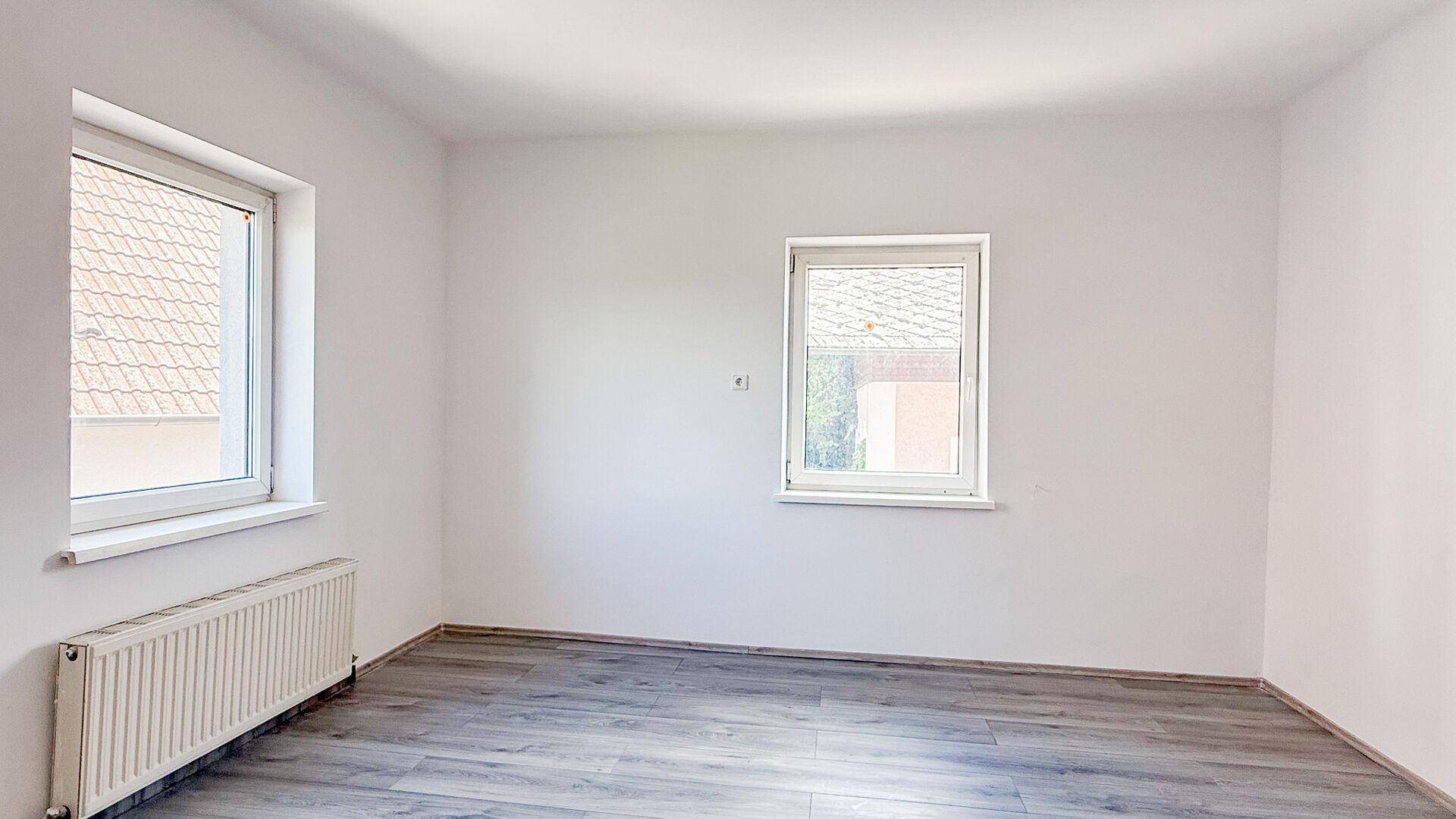 Leerer Raum mit weiß gestrichenen Wänden, grauem Laminatboden und zwei Fenstern mit Blick auf Dächer.