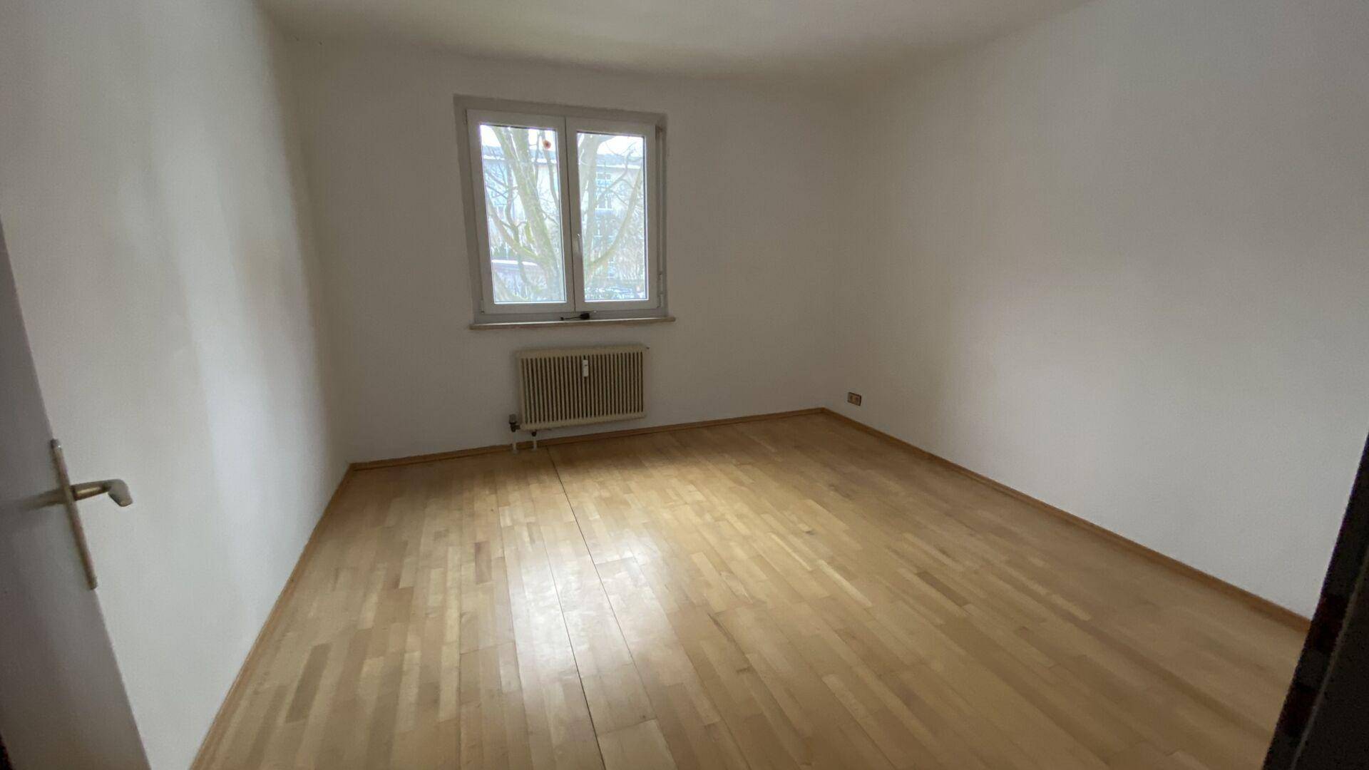 Schlafzimmer mit Parkettboden, weißen Wänden und einem Fenster mit Blick nach draußen.