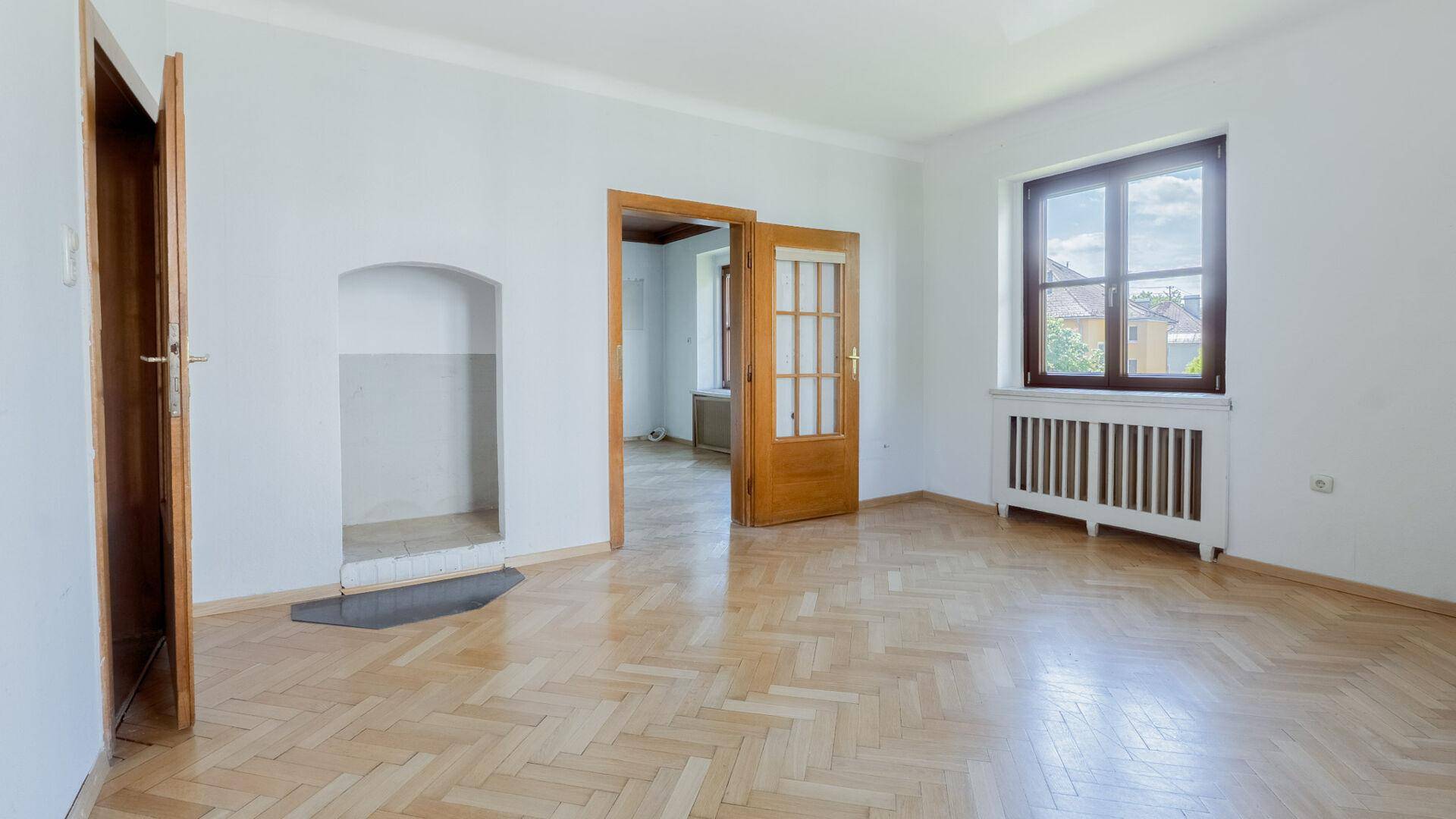 Leeres Wohnzimmer mit Fischgrätparkett, weiß gestrichenen Wänden und Kaminvorbereitung.