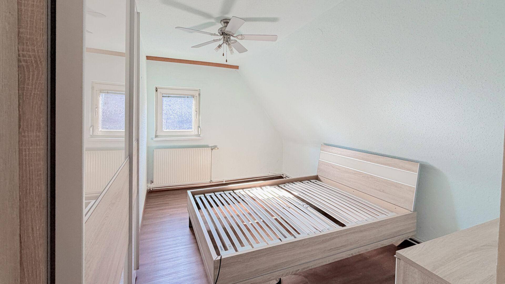 Schlafzimmer im Dachgeschoss mit Bettrahmen, Kleiderschrank und Deckenventilator.