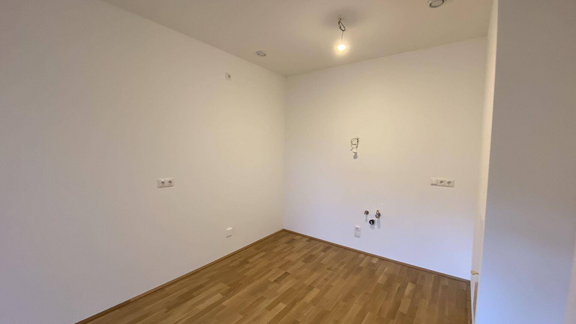 Leerer Küchenraum mit Parkettboden und Anschlüssen für eine Einbauküche.