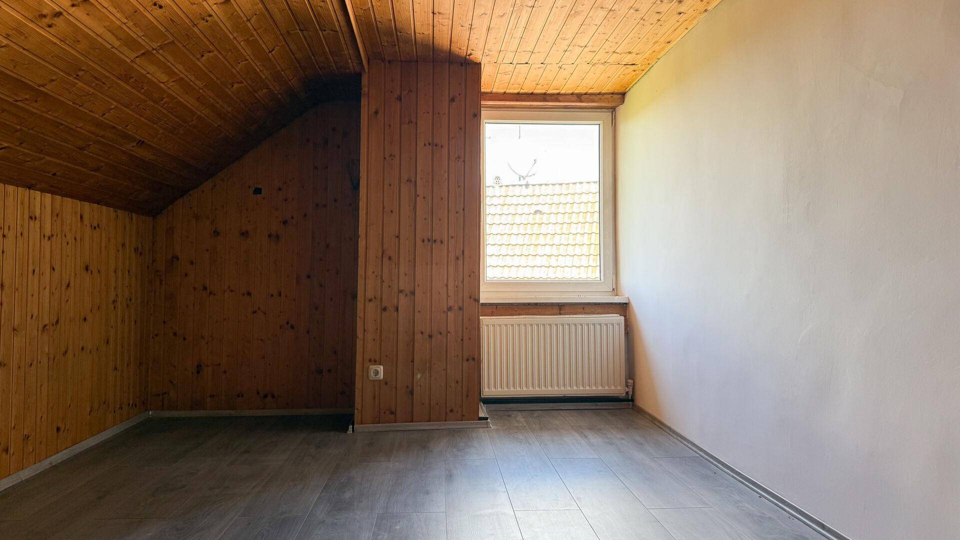 Helles Dachzimmer mit Holzverkleidung, schräger Decke und einem Fenster mit Blick nach draußen.