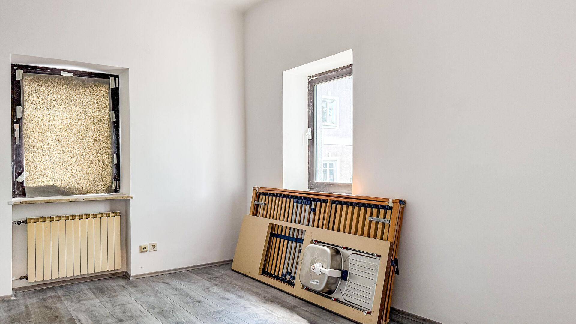 Leerer Raum mit zwei Fenstern, Heizkörper und einem zerlegten Küchenelement an der Wand.
