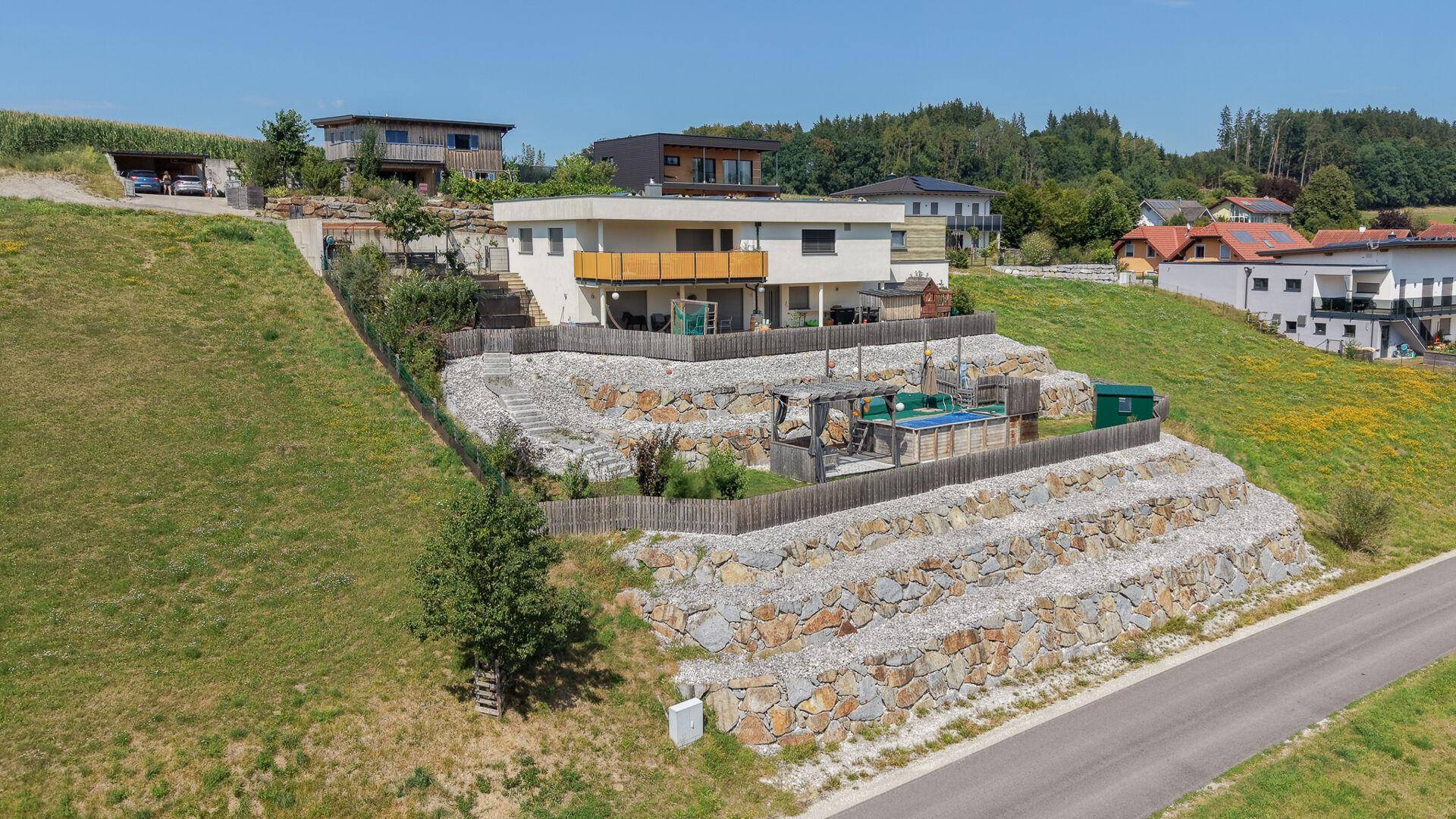 Modernes Haus am Hang mit terrassierter Landschaftsgestaltung und Balkon, umgeben von grüner Natur.