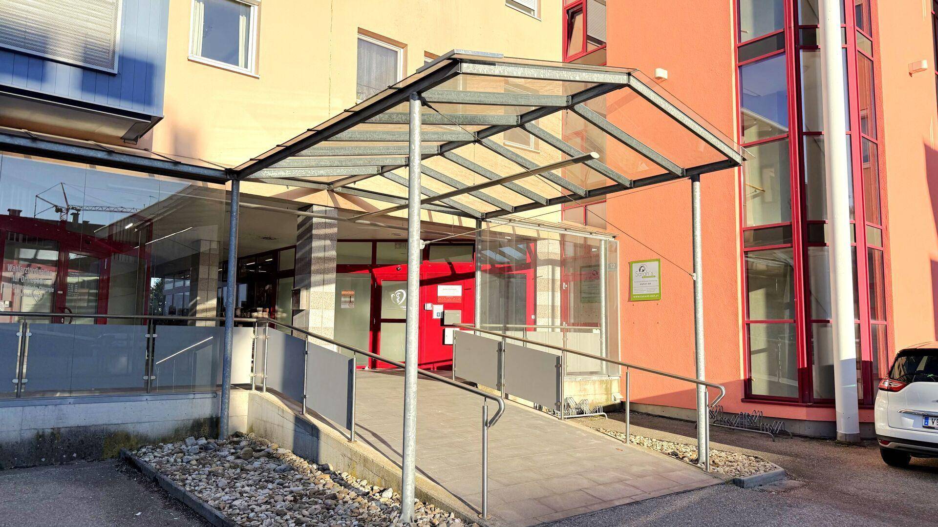 Gebäudeeingang mit Glasüberdachung und einer barrierefreien Rampe für Rollstuhlfahrer.