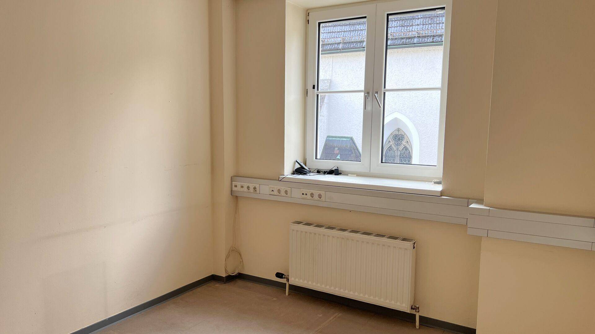 Kleiner, leerer Raum mit einem Fenster, das einen Blick auf ein Kirchengebäude im gotischen Stil bietet.