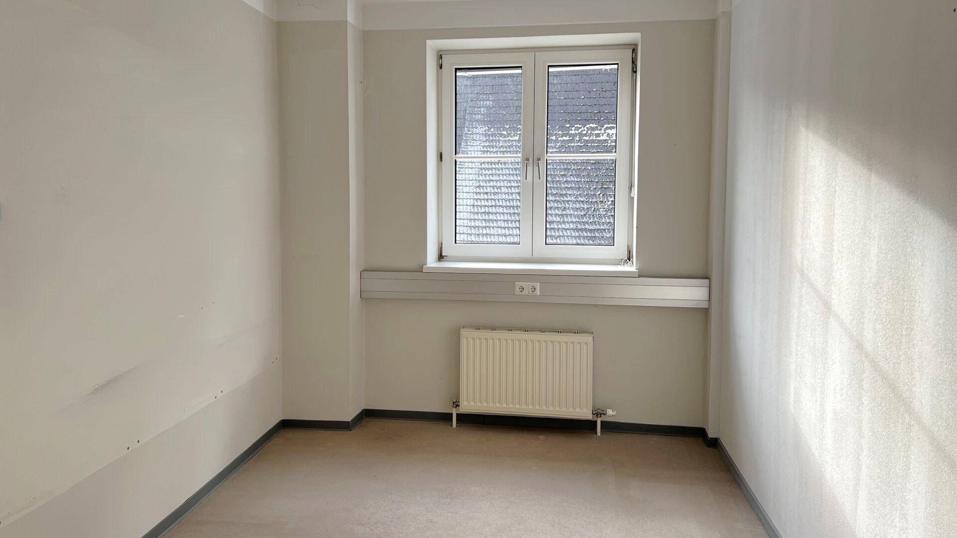 Heller, leerer Raum mit einem Fenster und einem Heizkörper, der vielseitig genutzt werden kann.