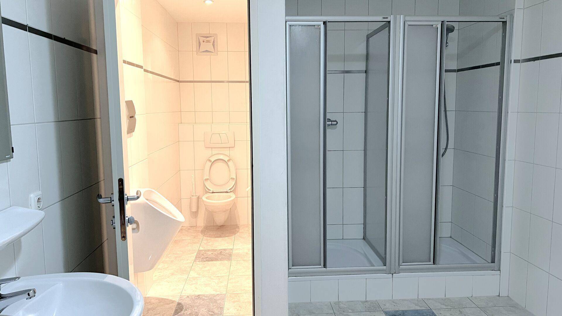 Funktionales Badezimmer mit Duschkabine, Toilette und Urinal, komplett gefliest in hellen Farben.