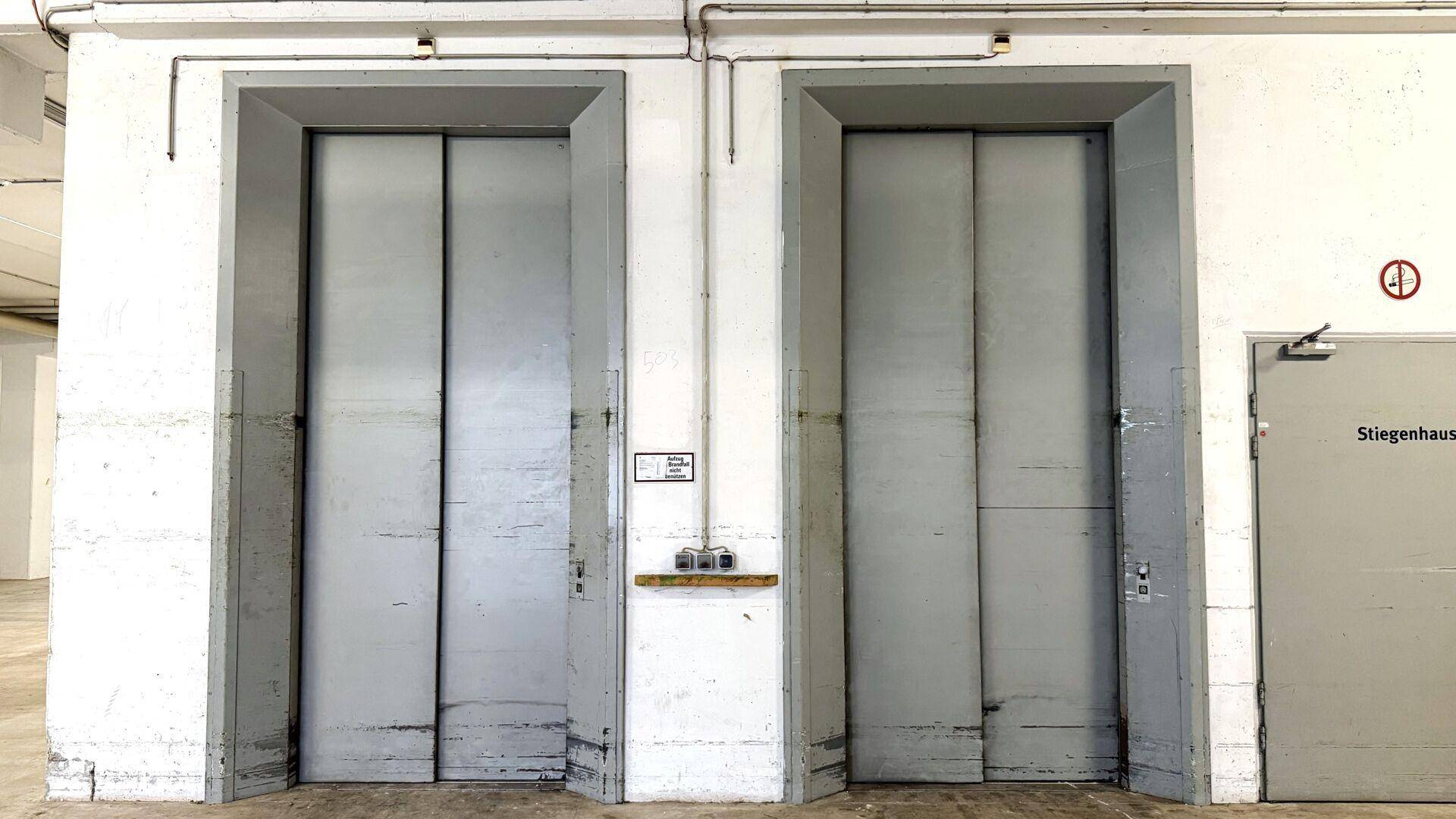 Zwei große Lastenaufzüge in einem Flur mit Betonwänden und einer Tür zum Treppenhaus.