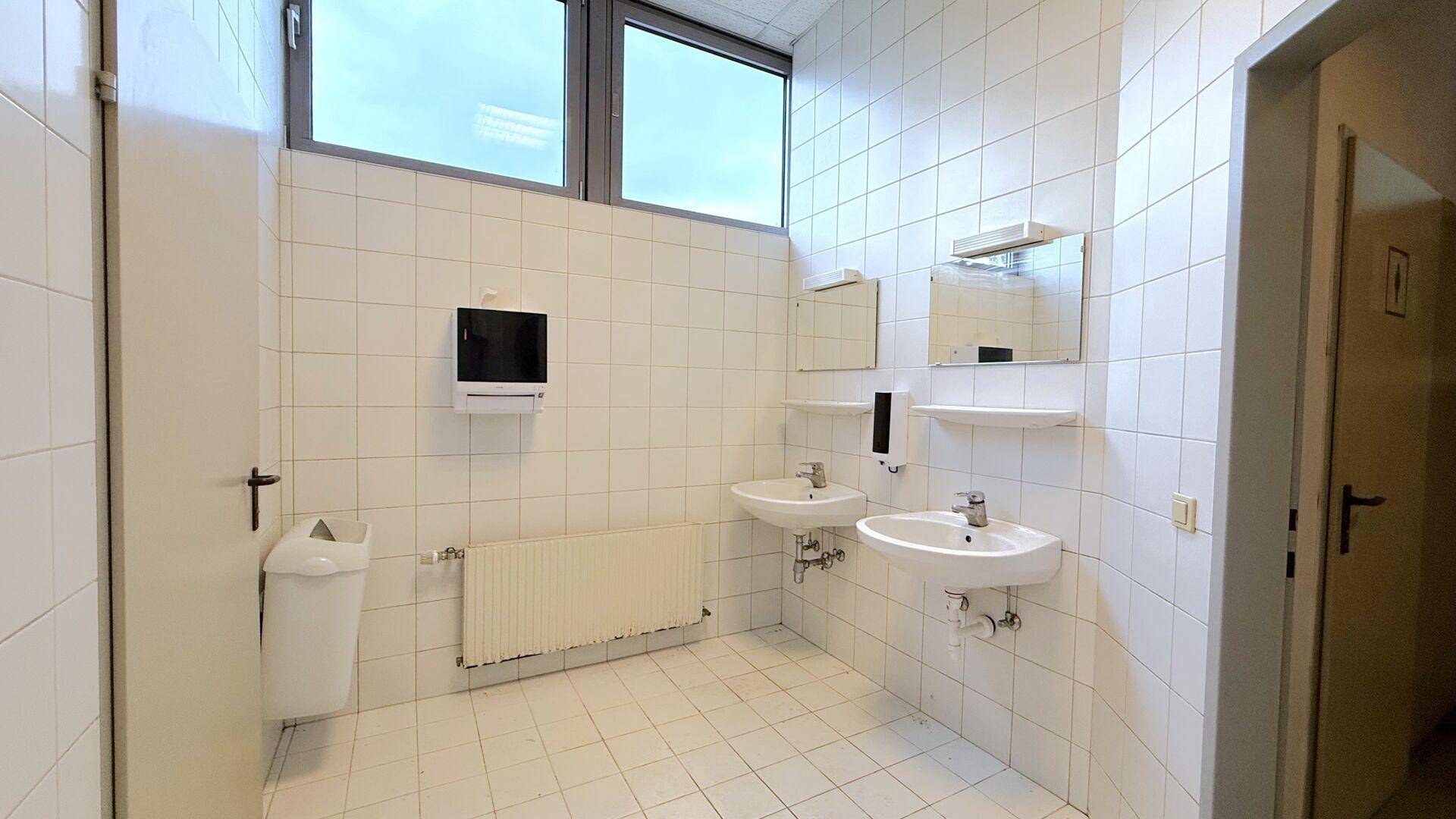 Heller Waschraum mit weißen Fliesen, zwei Waschbecken und einem großen Fenster.