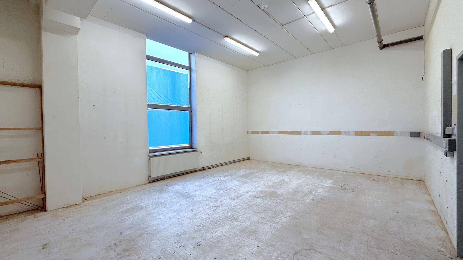 Kleiner, leerer Raum mit Betonboden, weiß gestrichenen Wänden und einem großen Fenster.
