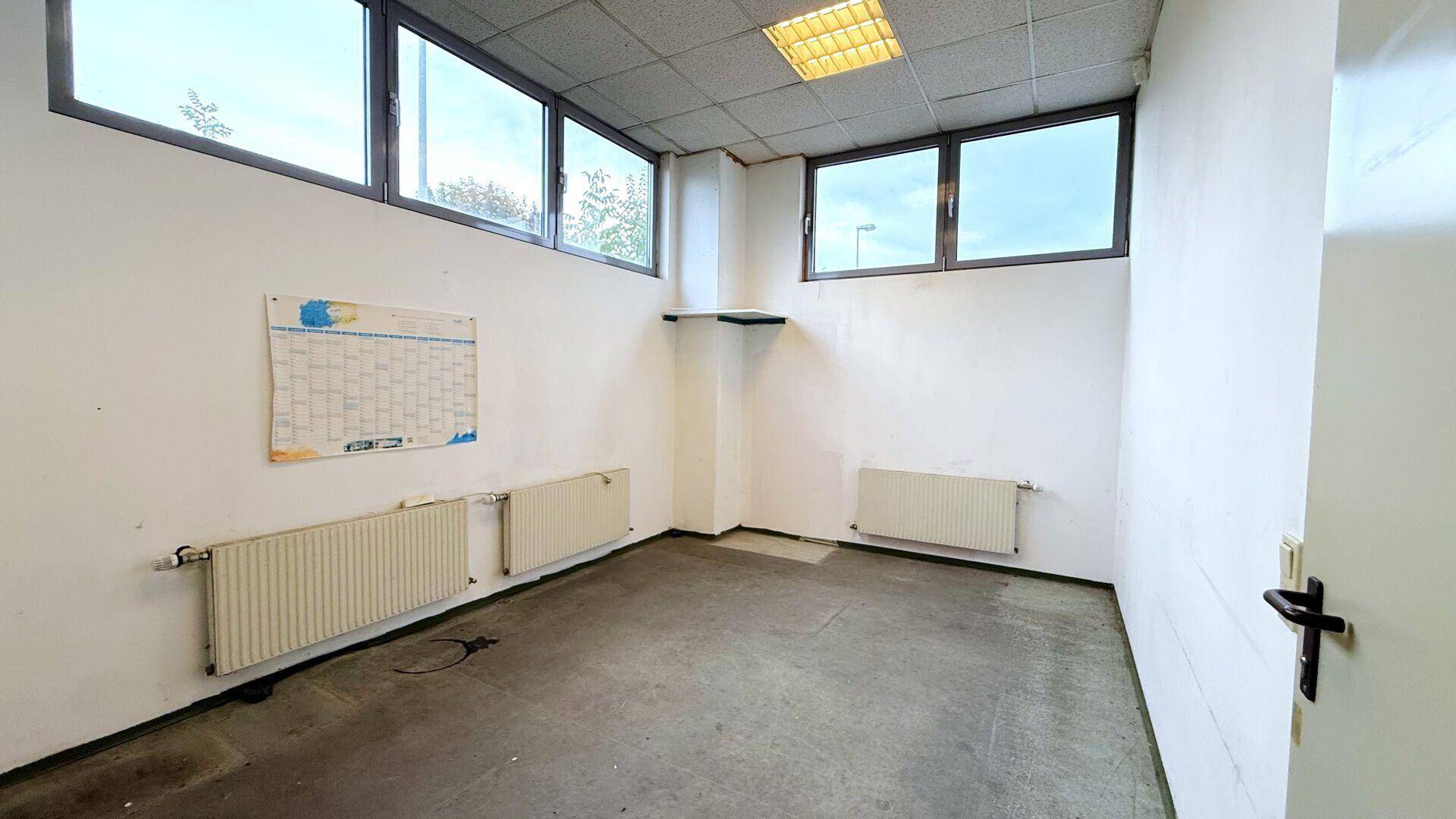 Kleiner Büroraum mit Fenstern, Heizkörpern und einem Kalender an der Wand.