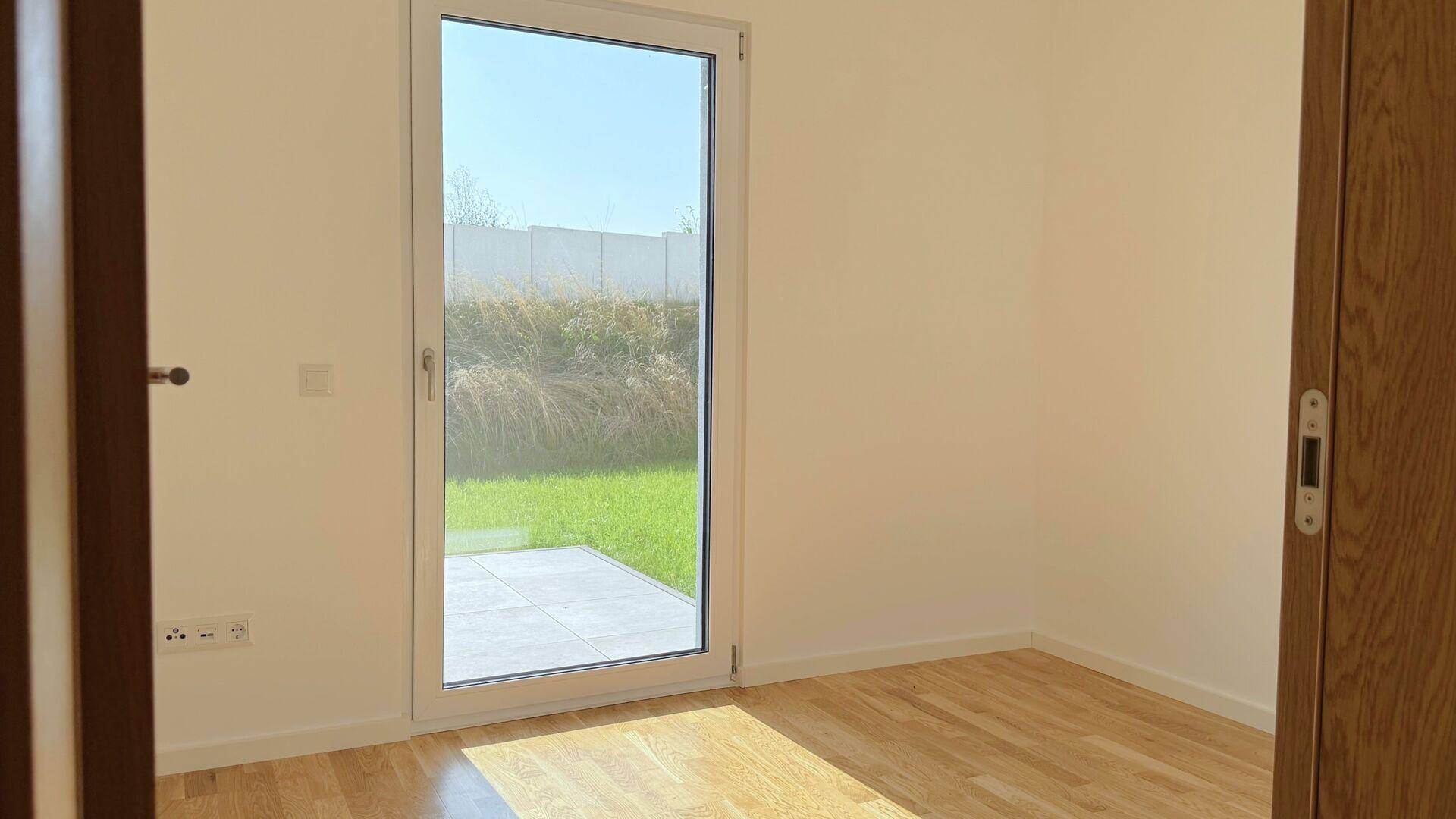 Helles Zimmer mit Holzboden und einer Glastür, die zu einem Außenbereich führt.