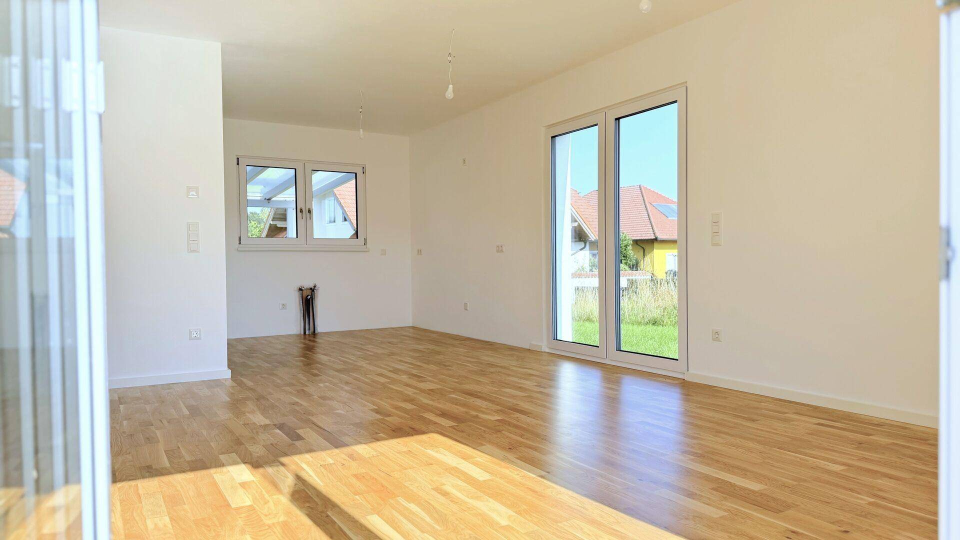 Helles, leeres Zimmer mit Holzboden und großen Fenstern, die viel Tageslicht hereinlassen.