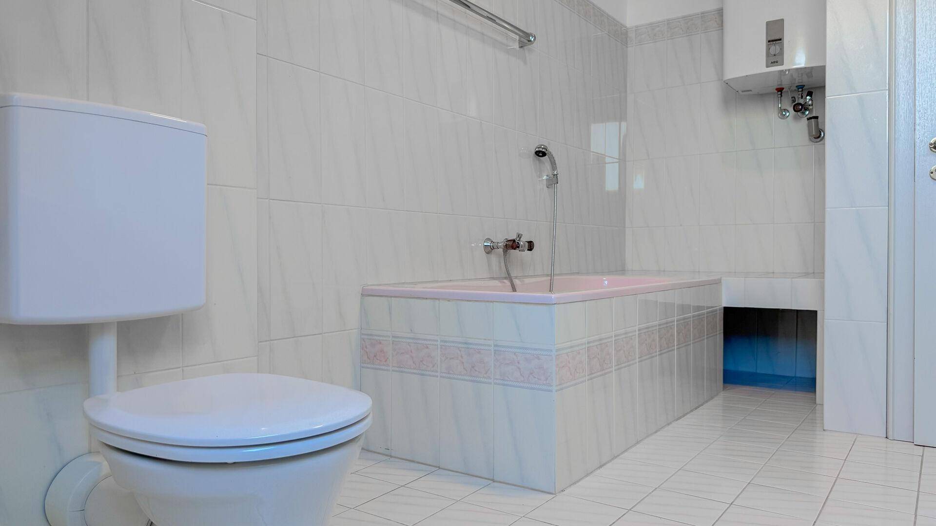 Badezimmer mit weißen Fliesen, Toilette und einer eingebauten Badewanne mit rosa Akzenten.