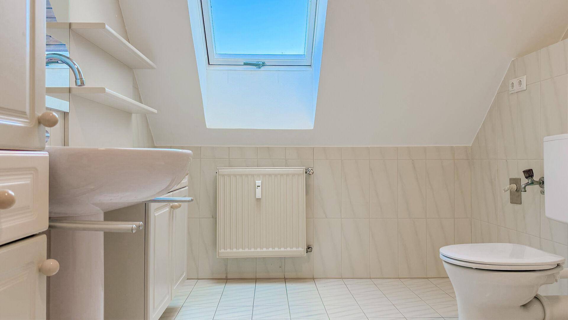 Helles Badezimmer mit weißen Fliesen, Waschtisch und Toilette unter einem Dachfenster.