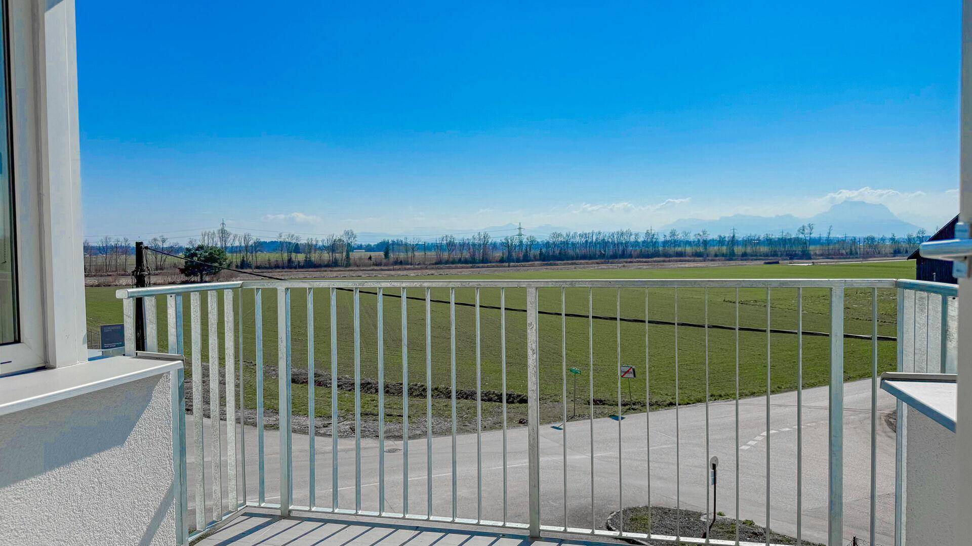 Sonniger Balkon mit Metallgeländer und weitem Blick über grüne Felder bis zu den Bergen.