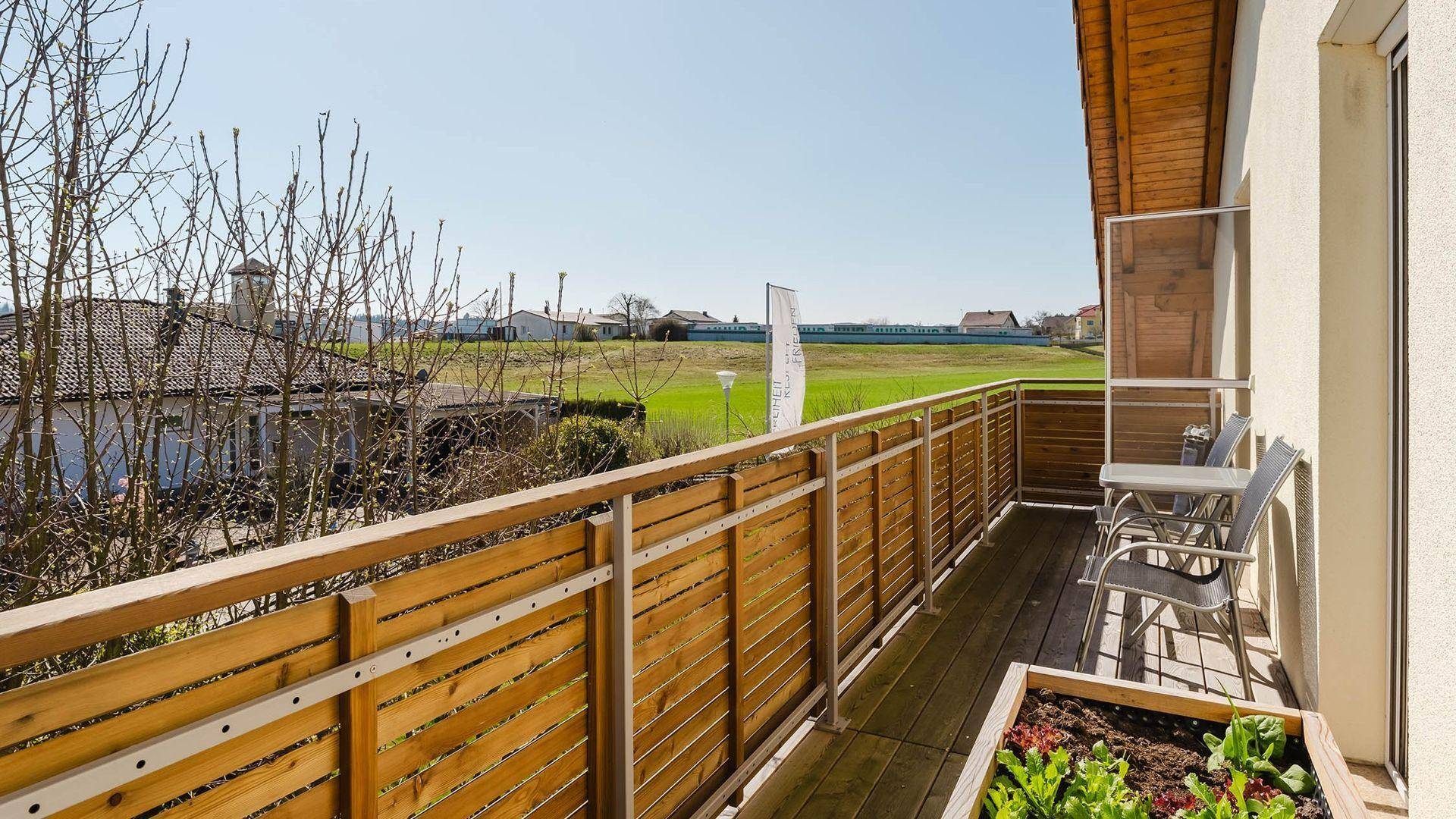 Sonniger Balkon mit Holzboden, Sitzgelegenheiten und weitem Blick über grüne Felder.