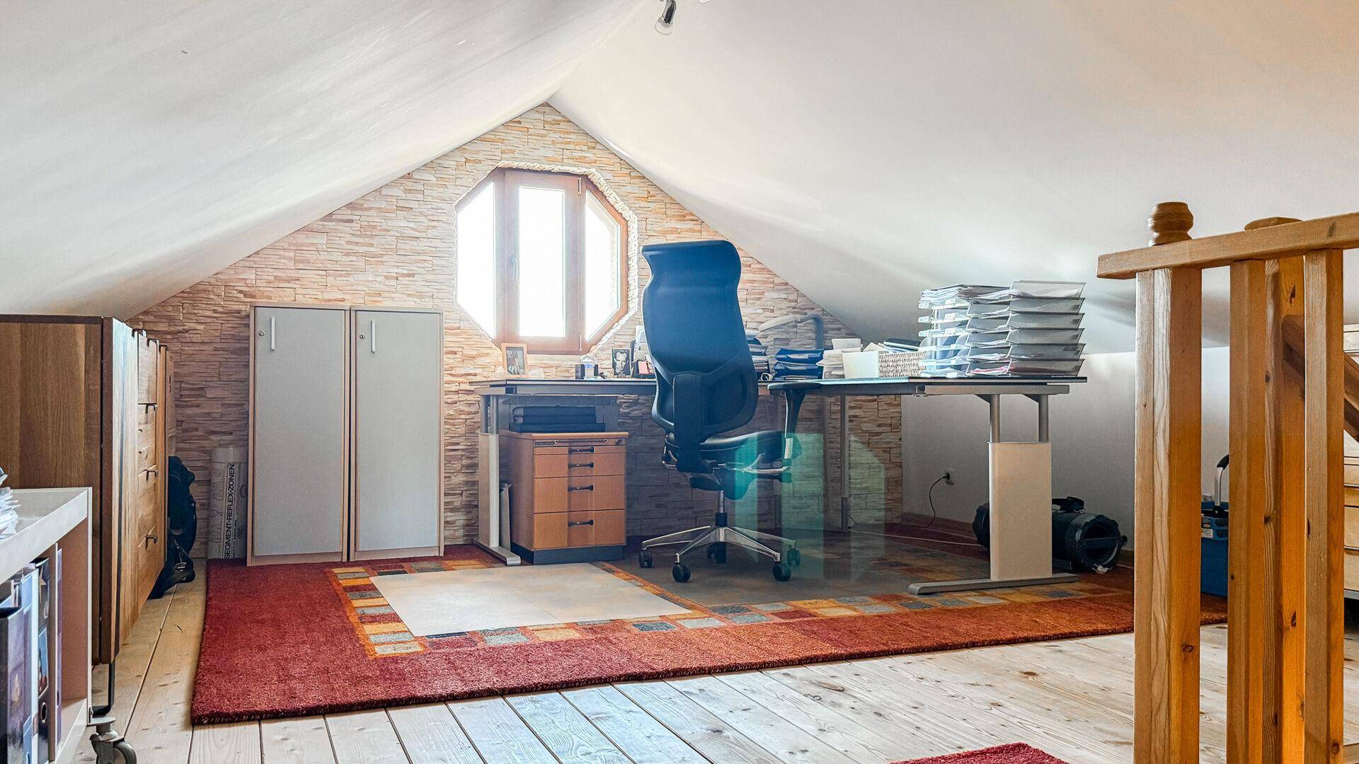 Geräumiges Dachgeschossbüro mit schräger Decke, großem Schreibtisch und besonderem Fenster.