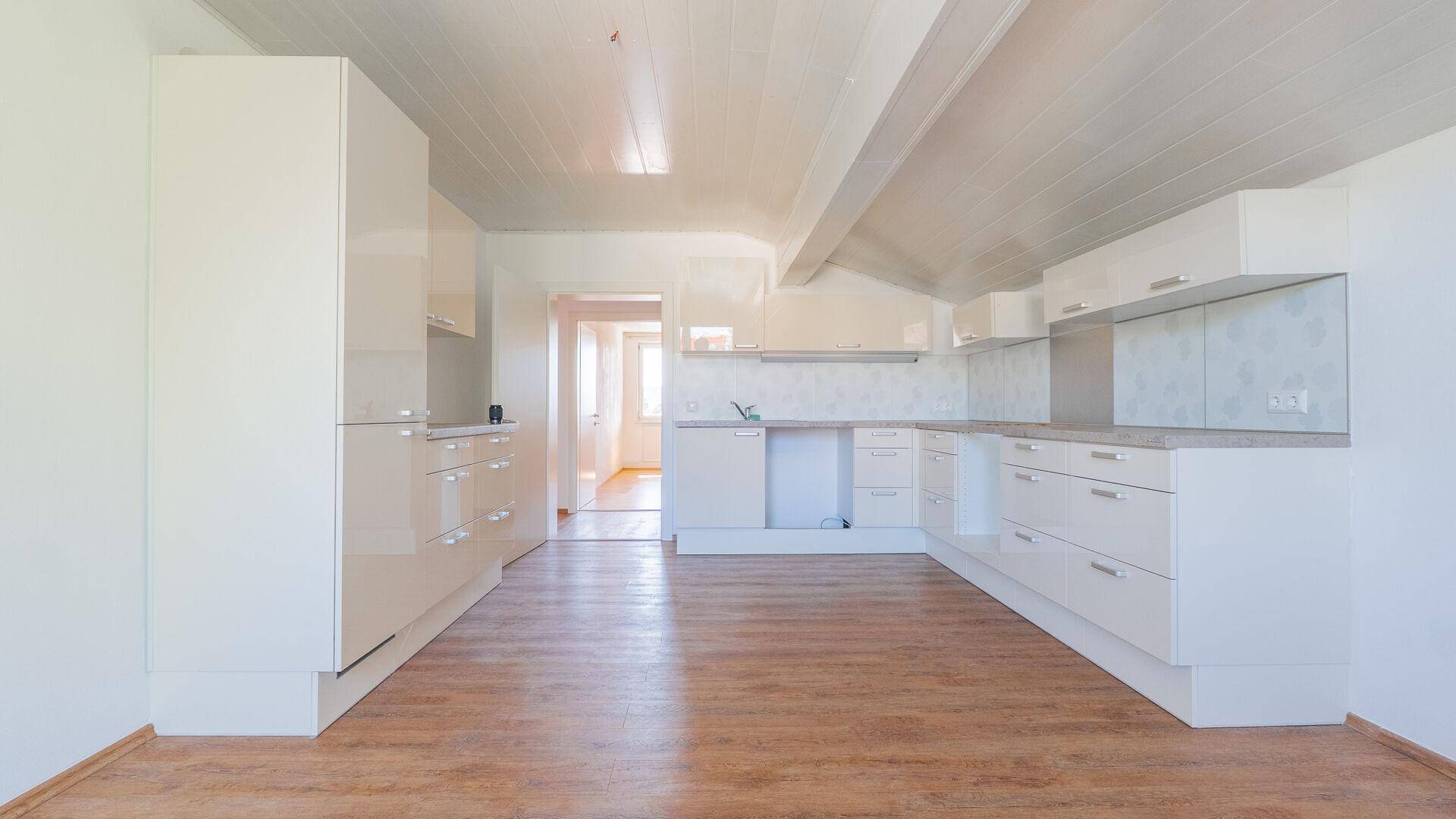 Geräumige, leere Küche mit weißen Schränken, Laminatboden und viel Tageslicht.