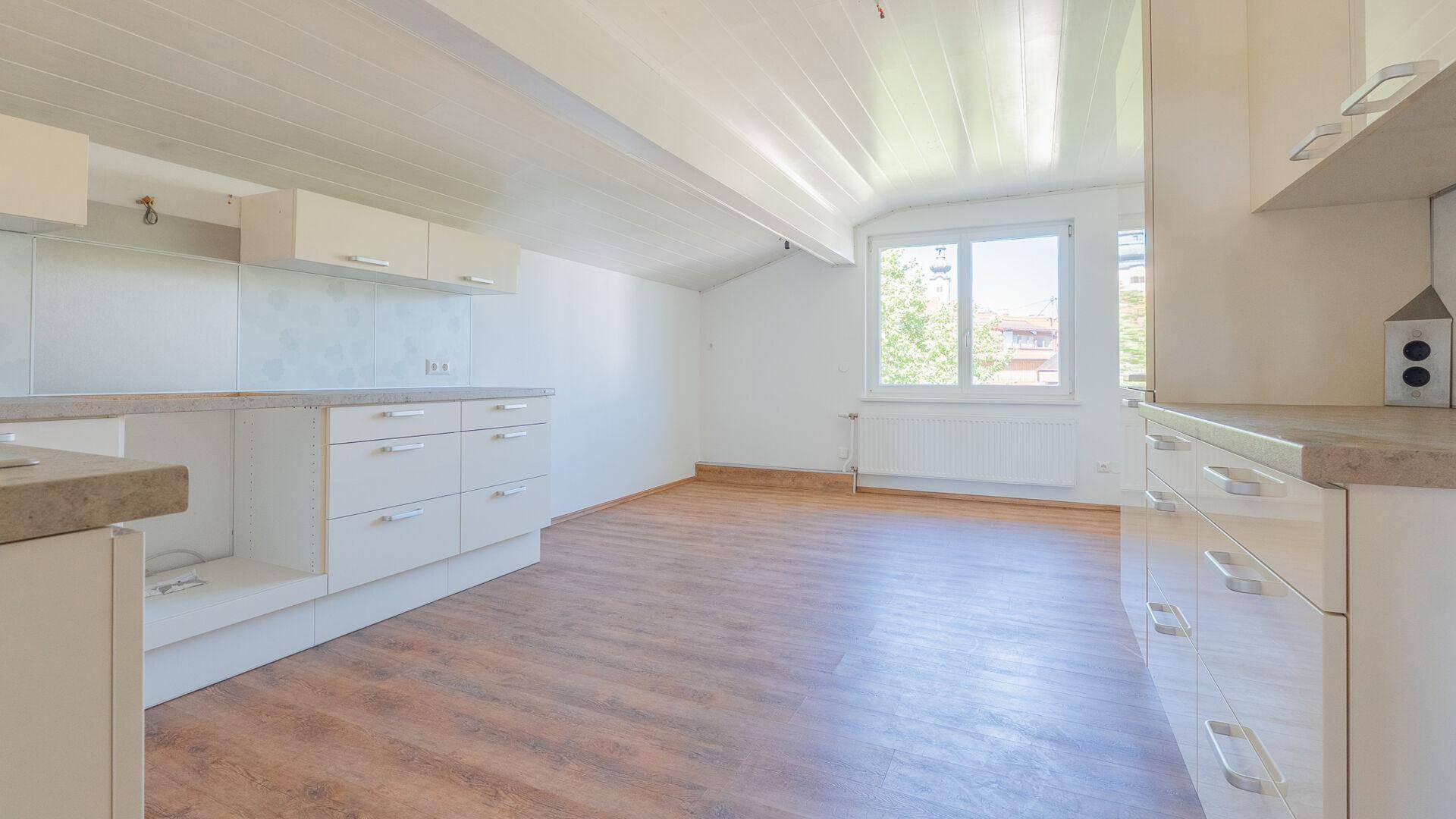 Leere Küchenzeile mit weißen Schränken, Arbeitsplatten und schräger Decke im Dachgeschoss.