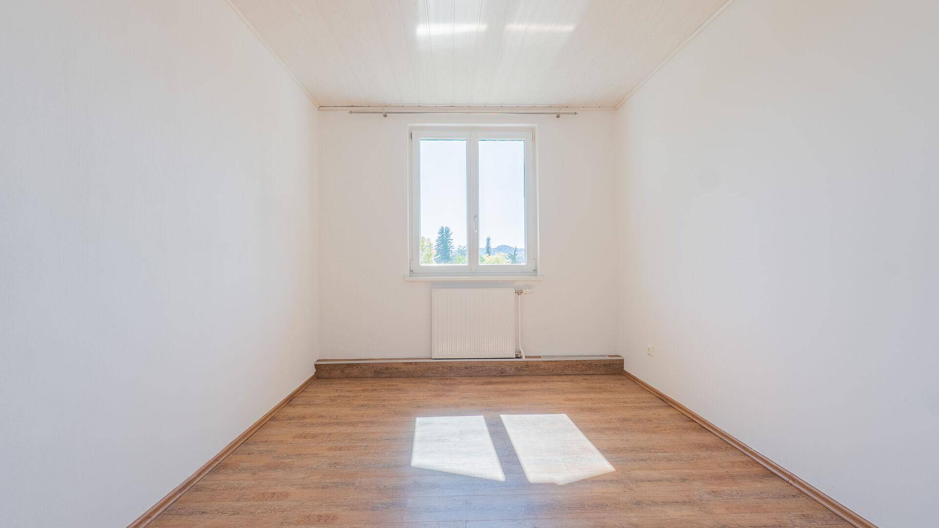 Leerer, heller Raum mit Laminatboden und einem Fenster, das viel Tageslicht hereinlässt.
