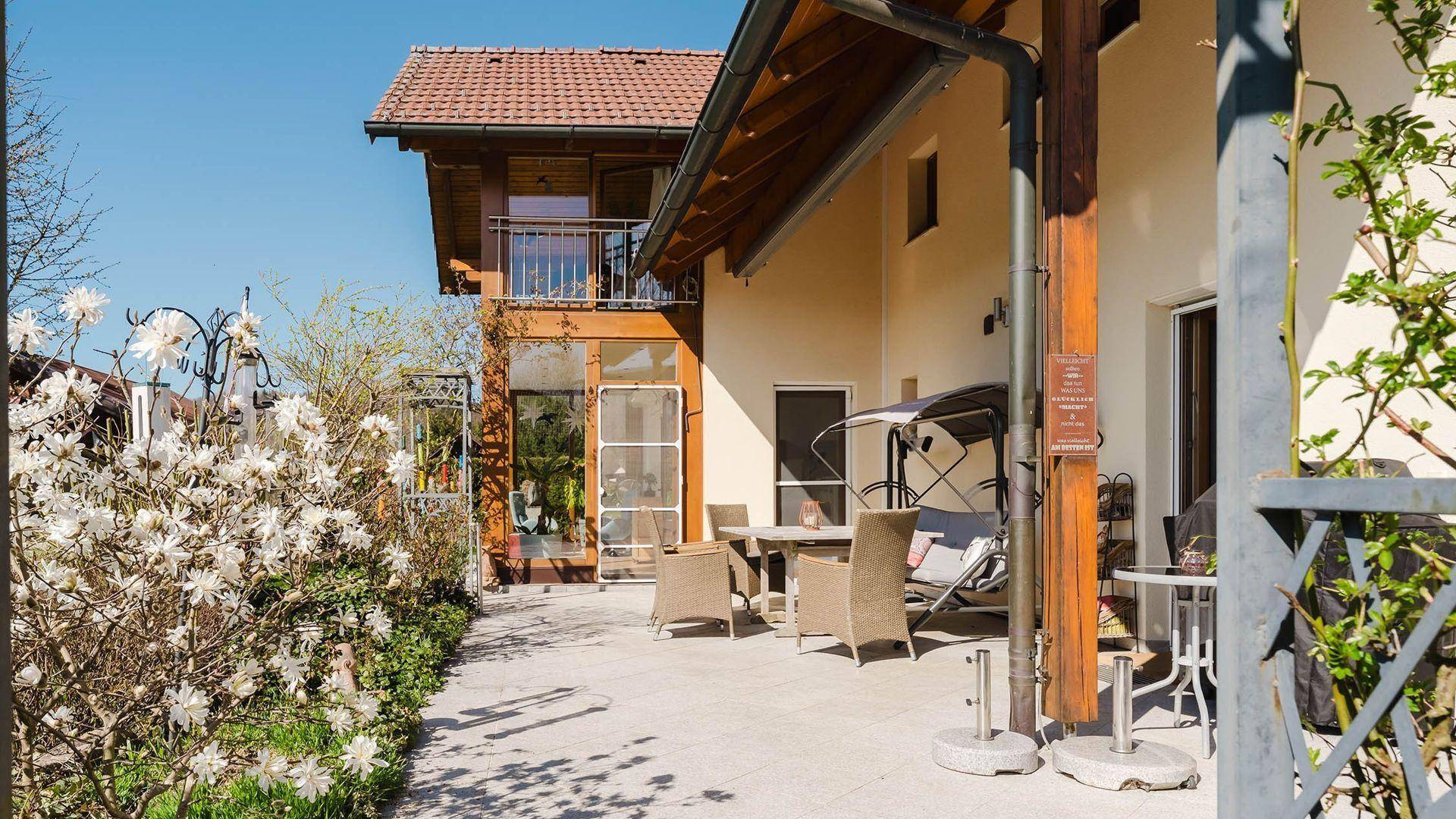 Sonniger Terrassenbereich mit Gartenmöbeln und blühenden Sträuchern neben dem Haus.