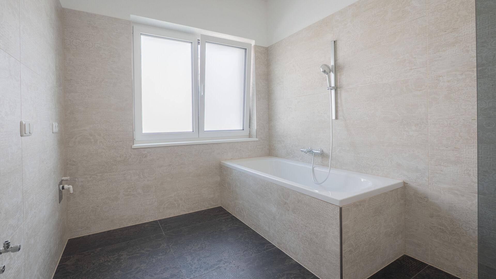 Helles Badezimmer mit Badewanne, hellen Wandfliesen und einem Fenster für natürliche Belüftung.
