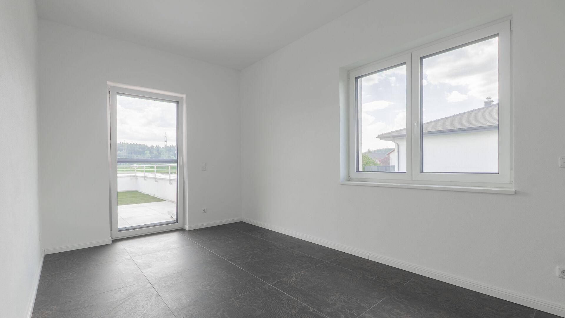 Heller Raum mit dunklem Fliesenboden, einem Fenster und Zugang zu einem Außenbereich.