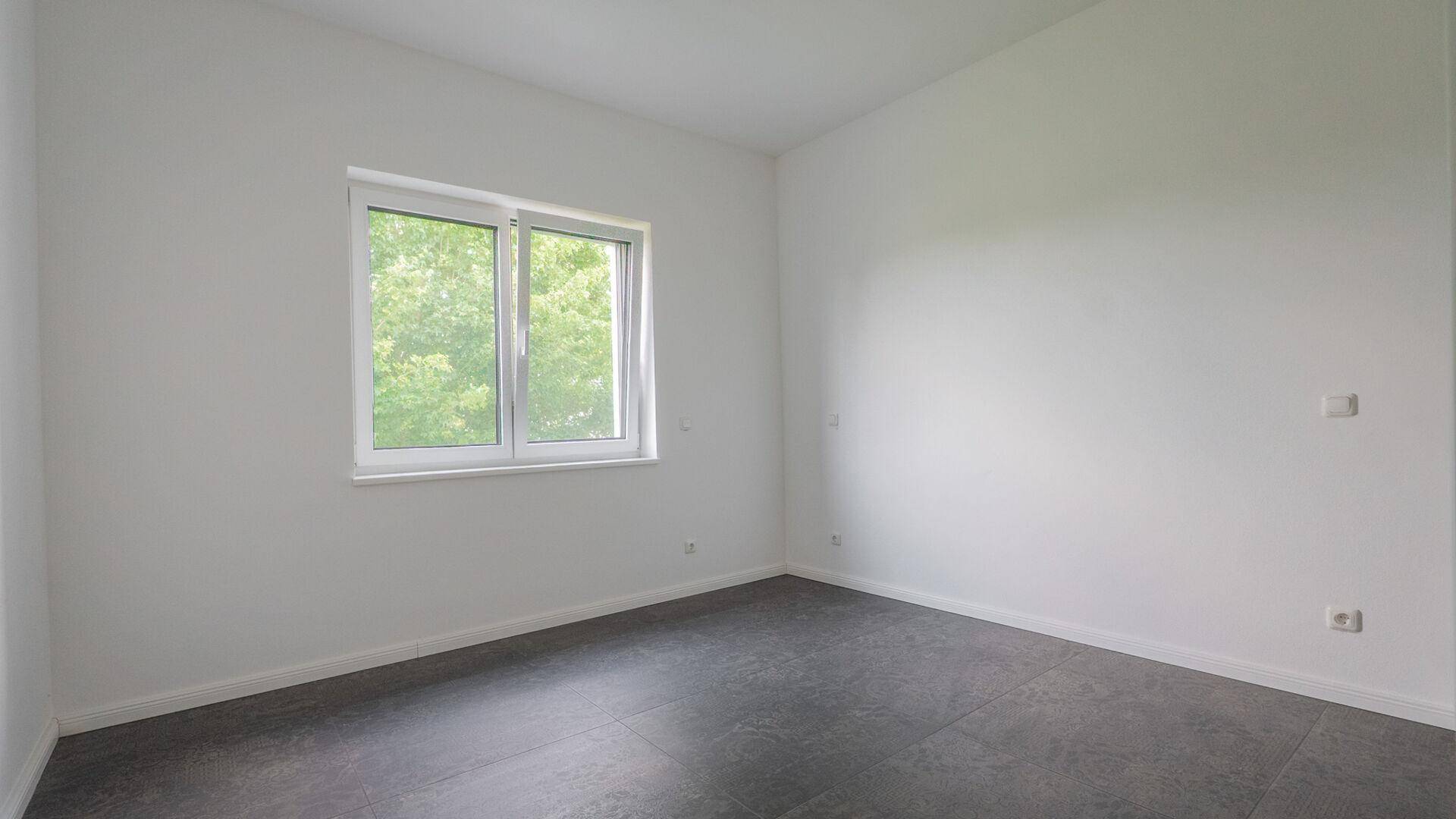 Leerer Raum mit dunklem Fliesenboden und einem Fenster mit Blick ins Grüne.