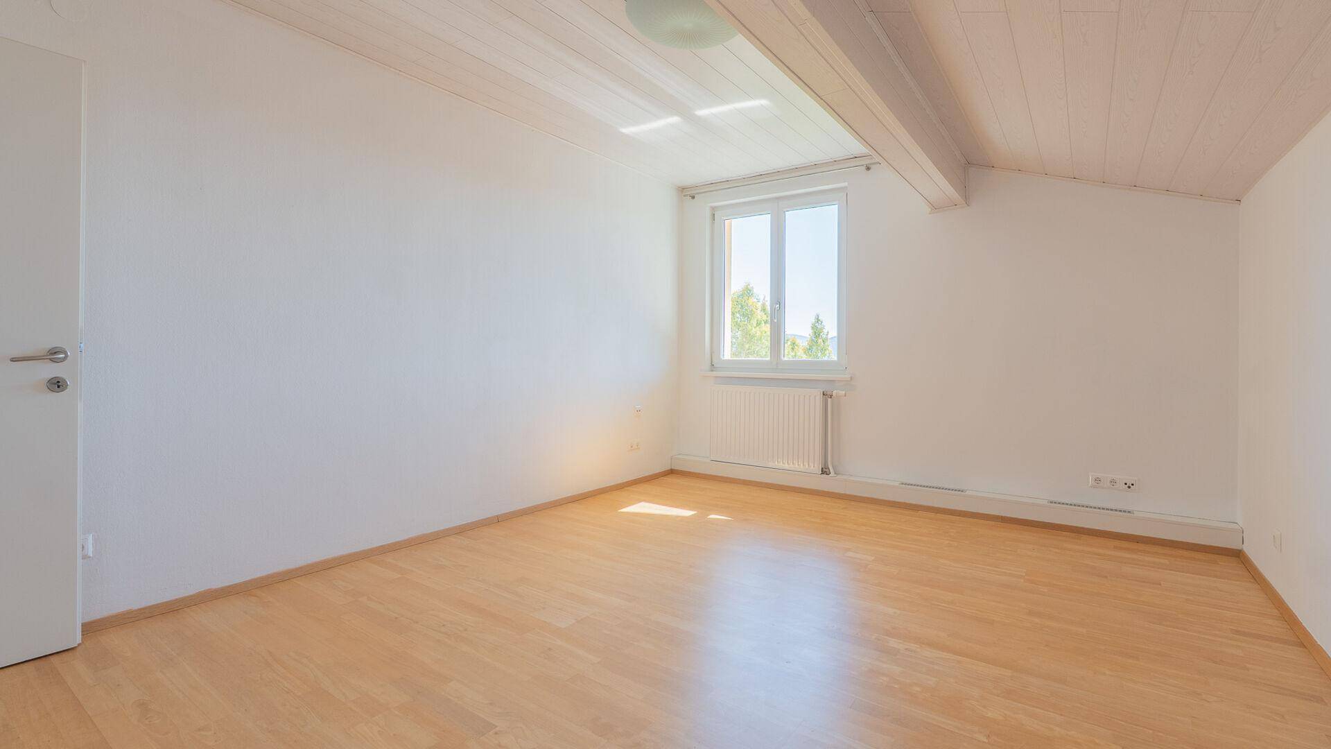 Heller Raum im Dachgeschoss mit Holzbalken, Laminatboden und einem großen Fenster.